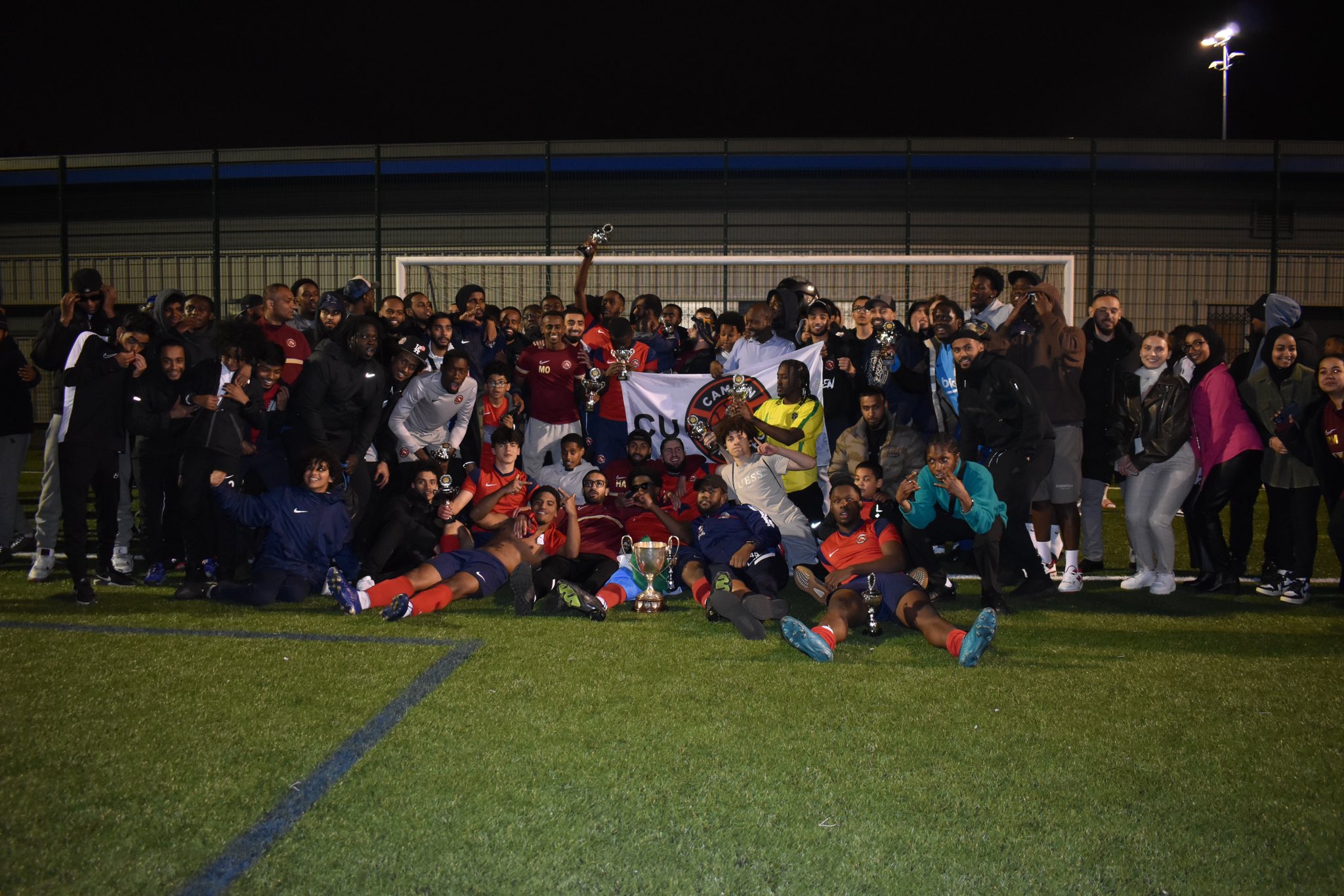 Camden United and Munye League players celebrating together on the pitch with trophies
